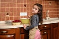 Charming child, little preschooler girl in chef's apron, cooks cherry pie in home kitchen, looks at camera over