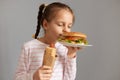 Charming cute beautiful little girl with pigtails with hot dog smelling aromat fresh baked hamburger standing with closed eyes Royalty Free Stock Photo