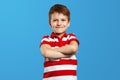 Charming cute kid in red stripped shirt crossing arms while standing against blue background