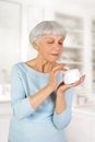 Charming elderly woman applying cosmetic cream on her face for facial skin care in bathroom at home.