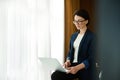 Charming elegant young prosperous pensive entrepreneur, business woman typing on a laptop a progress report, sitting in the edge