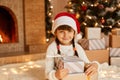 Charming female kid holding stack of present, little kid wearing white sweater and santa claus hat, sitting on floor near Royalty Free Stock Photo