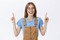 Charming friendly-looking charismatic female in glasses and trendy brown overalls pointing up with raised arms and