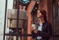 A charming girl wearing a leather jacket with a rucksack holding cup with takeaway coffee outside near the cafe. Royalty Free Stock Photo