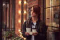 A charming girl wearing a leather jacket with a rucksack holding cup with takeaway coffee outside near the cafe. Royalty Free Stock Photo