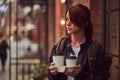 A charming girl wearing a leather jacket with a rucksack holding cup with takeaway coffee outside near the cafe. Royalty Free Stock Photo