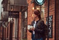 A charming girl wearing a leather jacket with a rucksack holding cup with takeaway coffee outside near the cafe. Royalty Free Stock Photo