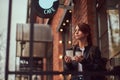 A charming girl wearing a leather jacket with a rucksack holding cup with takeaway coffee outside near the cafe. Royalty Free Stock Photo