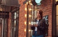 A charming girl wearing a leather jacket with a rucksack holding cup with takeaway coffee outside near the cafe. Royalty Free Stock Photo