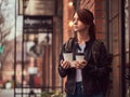 A charming girl wearing a leather jacket with a rucksack holding cup with takeaway coffee outside near the cafe. Royalty Free Stock Photo