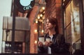 A charming girl wearing a leather jacket with a rucksack holding cup with takeaway coffee outside near the cafe. Royalty Free Stock Photo