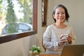 Happy 60s retired Asian woman holding a tea cup, smiling and looking at the camera Royalty Free Stock Photo