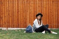Charming hipster girl with brown hair wearing a hat and backpack while sitting on the grass in park.