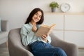 Charming Indian lady with cup of hot coffee and open book sitting in armchair at home Royalty Free Stock Photo