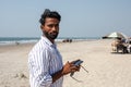 Charming Indian man is standing on beach against background of sea, holding phone in his hands and looking at camera. Royalty Free Stock Photo
