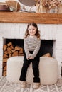 Charming little girl sits on soft pouffe chair relaxes at living room in Scandinavian style. Child in striped longsleeve