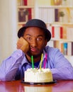 Charming man wearing blue shirt and hat sitting by table with birthday cake in front, looking sad depressed celebrating Royalty Free Stock Photo