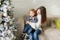 Charming motherin the white sweater holding her toddler son sitting on the sofa near Christmas tree in the house Royalty Free Stock Photo