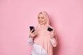 Charming Muslim woman in pink hijab, holding bank credit card and smartphone, makes payments via internet mobile banking Royalty Free Stock Photo