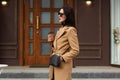 Charming mysterious woman stands outside, wears beige coat, leather black bag, fashionable sunglasses, holds her hand in pocket, Royalty Free Stock Photo