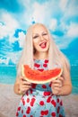 Charming plump blonde in blue pin up dress stands with a piece of watermelon and enjoys rest and a snack