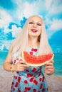 Charming plump blonde in blue pin up dress stands with a piece of watermelon and enjoys rest and a snack