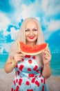 Charming plump blonde in blue pin up dress stands with a piece of watermelon and enjoys rest and a snack