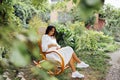 Charming pregnant brunette in elegant white dress is sitting on yellow rocking chair outdoors in garden