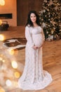 Charming pregnant brunette stands alone in the room and gently hugs her tummy with two hands, dressed in a long white Royalty Free Stock Photo