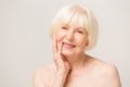 Charming, pretty, old woman touching her perfect soft face skin with fingers, smiling at camera over gray background, using day, Royalty Free Stock Photo