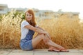 Charming red-haired girl in denim overalls sitting on a background of yellow dry grass at sunset in summer Royalty Free Stock Photo