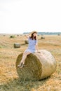 Charming red haired girl in hat and summer striped dress sitting on a hay bale in warm summer sunny day, wheat field on Royalty Free Stock Photo