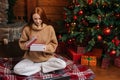 Charming redhead young woman opening Christmas gift box with present on background of xmas tree. Royalty Free Stock Photo