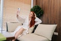 Charming redhead young woman taking selfie, having video call with friends using mobile phone, sitting on comfortable Royalty Free Stock Photo