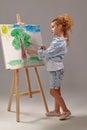 Charming school girl is painting with a watercolor brush on an easel, standing on a gray background.