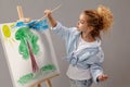 Charming school girl is painting with a watercolor brush on an easel, standing on a gray background.