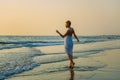 Charming slender blonde in stylish dress stands in waves on the sea. Young woman walks barefoot along the surf and enjoys the rest