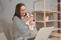 Charming smiling Caucasian woman wearing striped shirt sitting on sofa with her puppy dog using laptop computer for online Royalty Free Stock Photo
