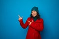 Charming smiling curly dark-haired woman in bright red coat and woolen green hat poses against blue background, looks at camera