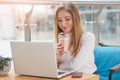 Charming smiling girl, wears casual sweater sits on sofa, drinks cocktail. Young woman watches film on laptop while resting or Royalty Free Stock Photo