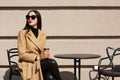 Charming stylish model with red lipstick wearing big trendy sunglasses, beige coat, black sweater and trousers, holding papercup