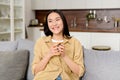 Charming teen lady holding mug of hot drink, looking away, daydreaming and holding coffee, sitting on the couch in a Royalty Free Stock Photo