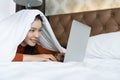 Charming teenage covering head with white blanket lying on bed and watching tv or movies streaming on laptop