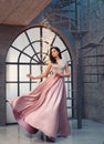 Charming tender princess in long peach pink shiny satin flying waving dress with neckline, girl stands on vintage stairs