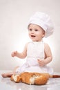 Charming toddler baby in hat of cook and apron sitting with bread loaves, laughing happily