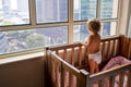 a charming toddler looks out the window at the metropolis while standing in a crib. baby in a crib looks out the window