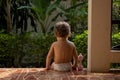 A charming toddler sits on the steps of a house in the sunlight. back view