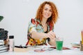 Charming woman with curly ginger hair drawing with a brush, sitting behind desk