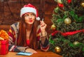 Charming Woman in a Red Hat with White Fur Sits at a Table near a Decorated Christmas Tree and Thinks what to Write to Royalty Free Stock Photo