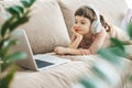 A charming 5-6-year-old girl lying on a couch, wearing headphones and watching a laptop screen. Concept: technology-infused